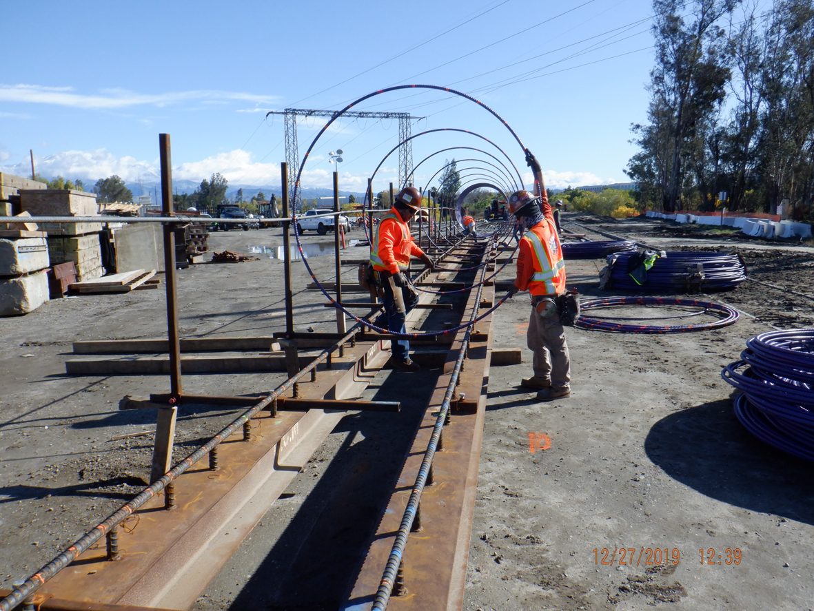 Two construction workers in safety gear assemble large metal reinforcement cages on a worksite under a clear sky.