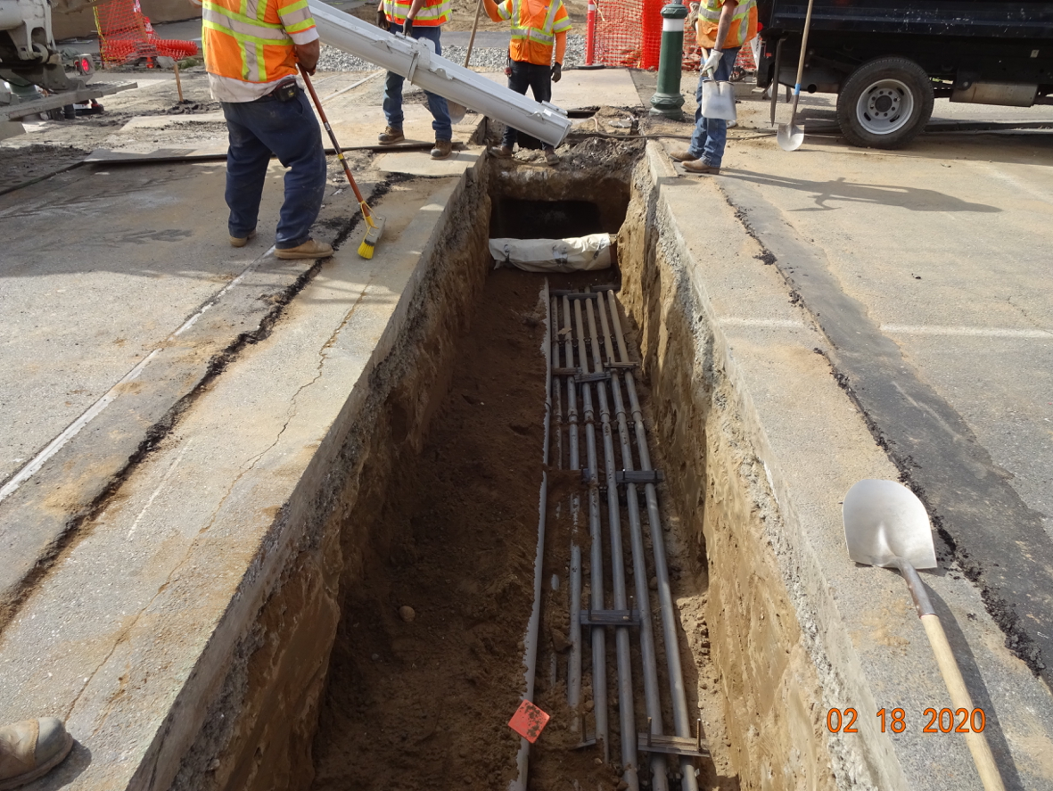 Workers in safety vests stand near an open trench with multiple utility conduits exposed underground at a construction site.
