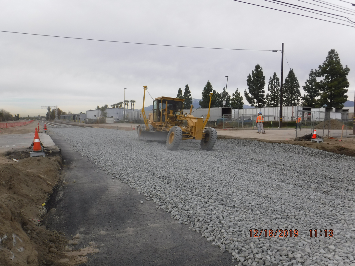 A road grader spreads gravel on a construction site while two workers stand nearby. Orange cones line the area; trees and buildings are in the background.