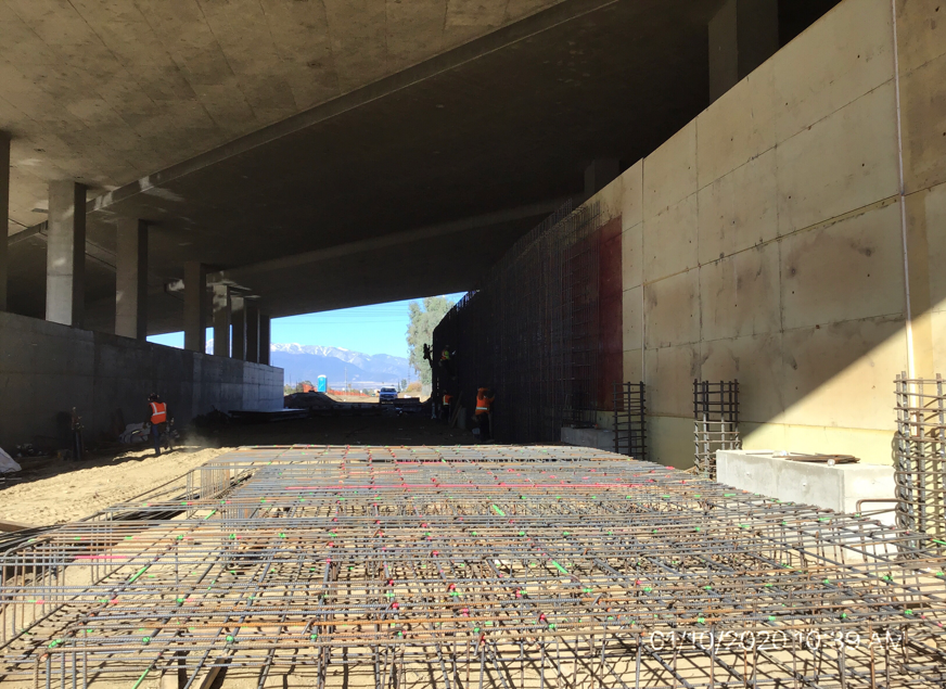 Construction workers install rebar under a large concrete overpass, with shadows cast by the structure and materials scattered on the ground.