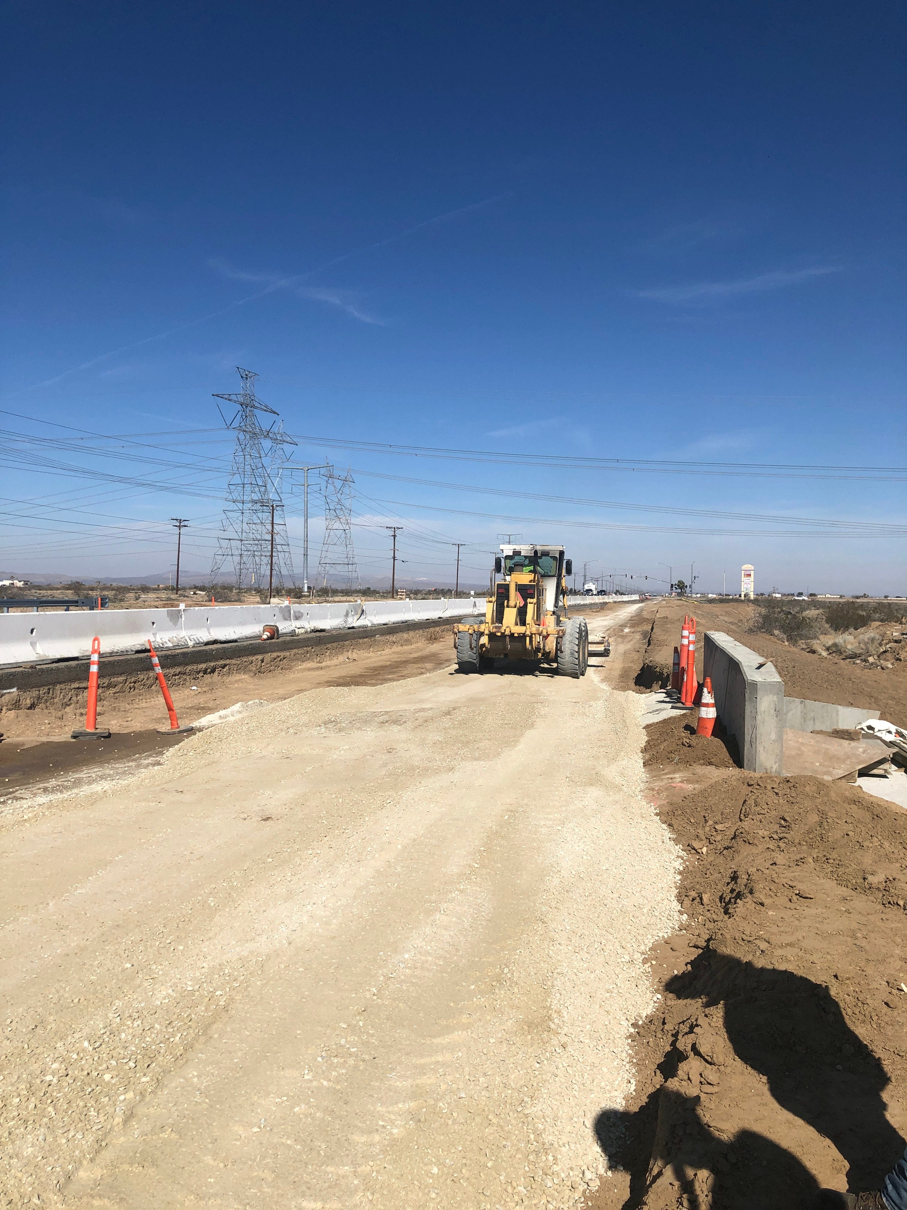 A construction grader works on a dirt road under clear blue sky, with orange traffic cones, power lines, and barriers visible nearby.
