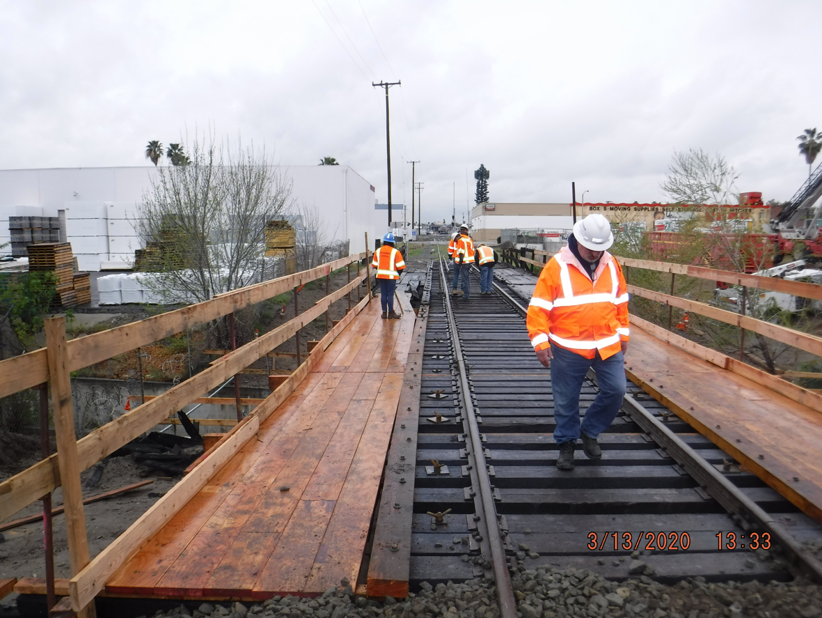 Workers in orange safety jackets and hard hats walk on a wooden walkway and railway tracks at a construction site on a cloudy day.