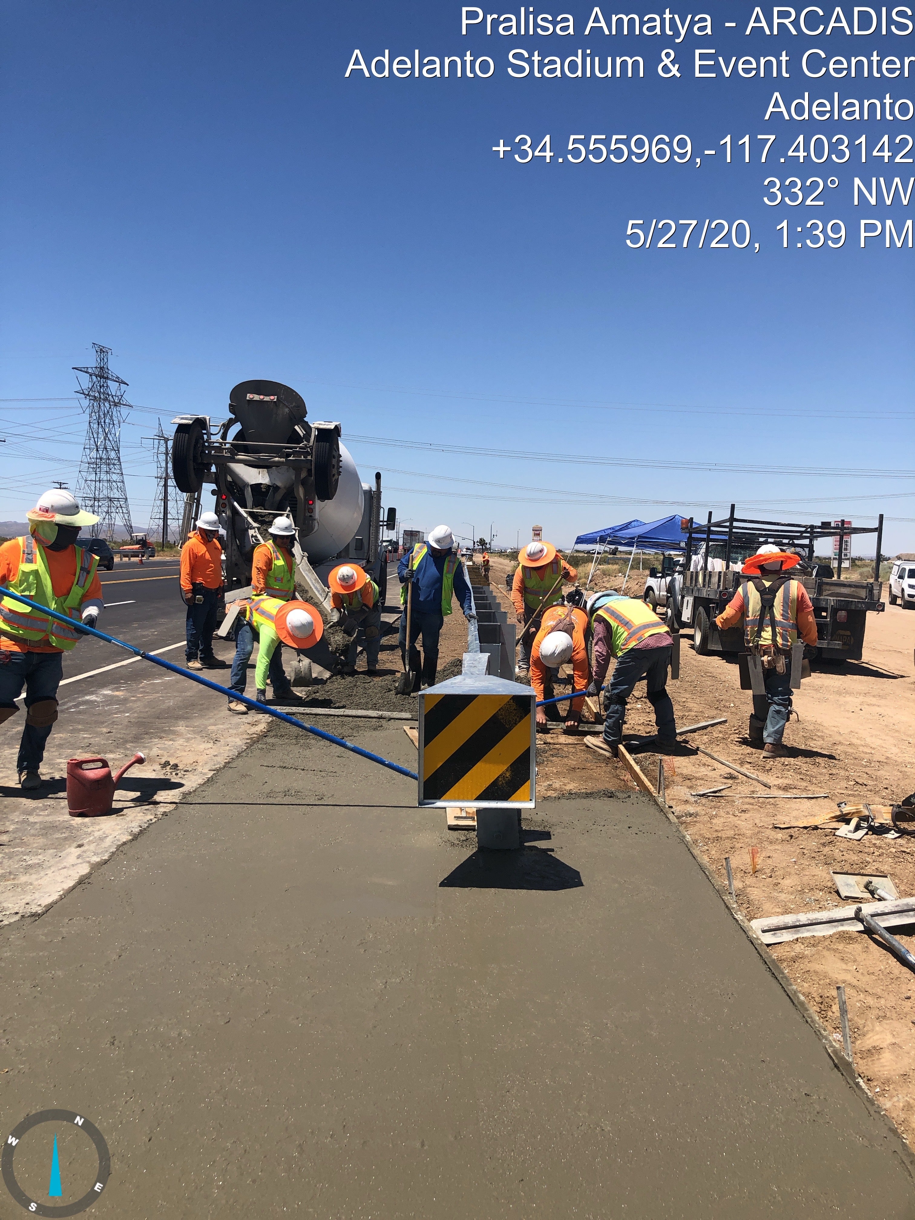 Construction workers pouring and smoothing concrete on a roadside project under clear skies, with a cement mixer truck and safety barriers visible. Text overlay provides project details and coordinates.