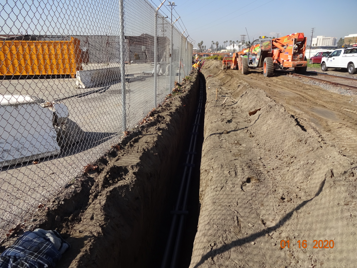 A long trench with exposed utility conduit runs parallel to a chain-link fence at a construction site, where workers and equipment are visible in the background.