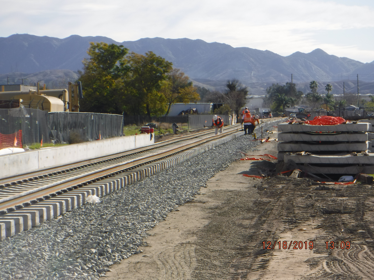 Construction workers walk along newly laid railway tracks with construction materials and equipment on the side, mountains visible in the background.