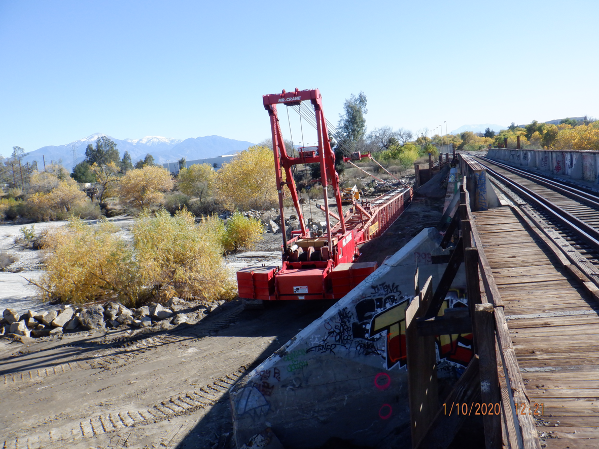 A red crane sits next to a railway bridge over a dry riverbed, with trees and mountains in the background and graffiti on a concrete wall in the foreground.