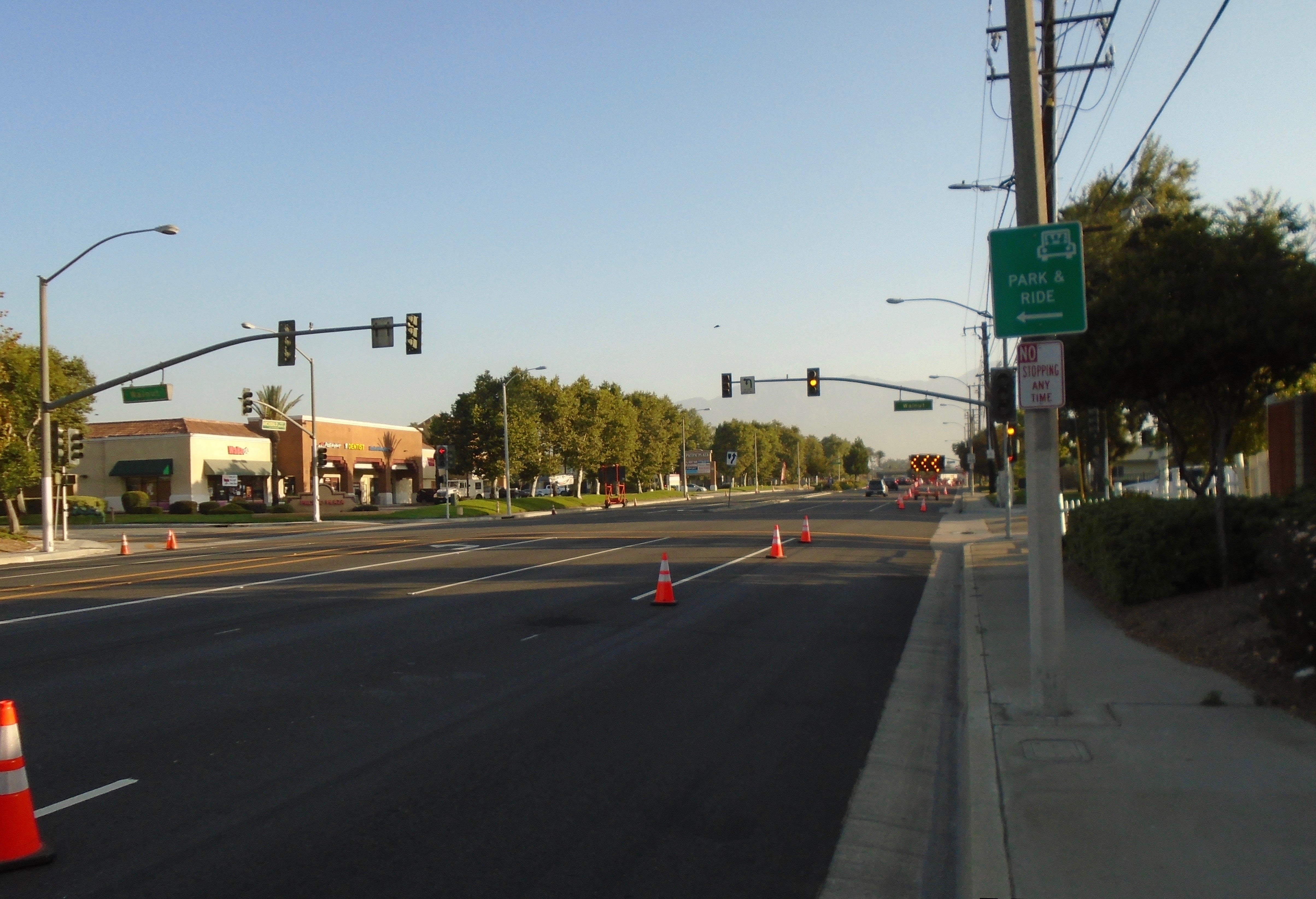 Wide street with traffic cones marking a closed lane, traffic lights ahead, businesses on the left, and a Park & Ride sign on the right sidewalk.