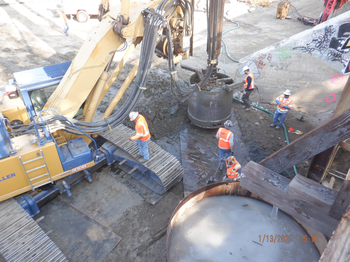 Construction workers in safety gear operate and monitor heavy machinery at an urban job site with concrete, dirt, and graffiti-covered walls visible.