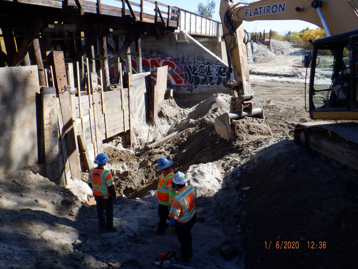 Three construction workers in safety vests and helmets stand near an excavator at a dirt worksite under a wooden bridge with graffiti.
