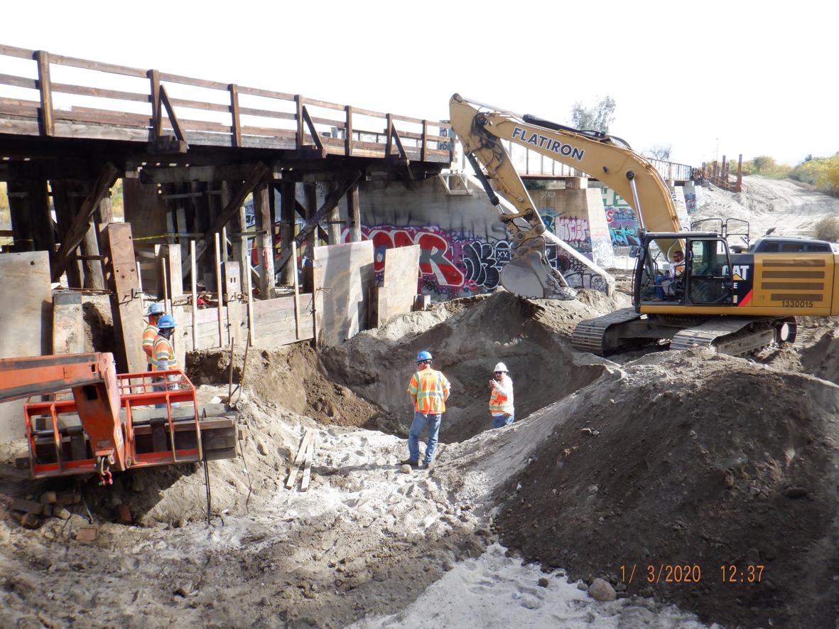 Construction workers and excavator operate near a wooden bridge with graffiti on its support pillars, preparing or repairing the bridge structure.
