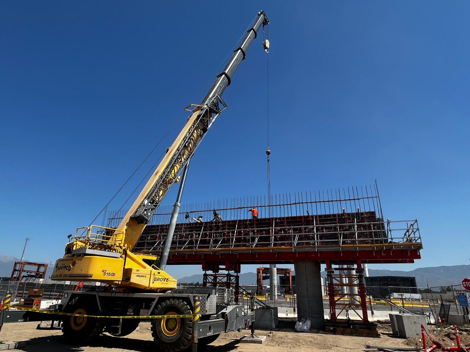 A large yellow crane lifts materials near a partially constructed bridge with workers standing on top, under a clear blue sky at a construction site.