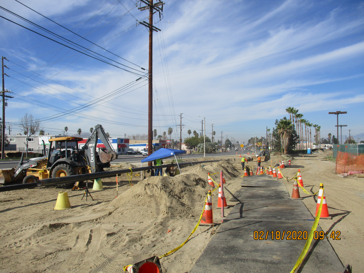 A construction site with traffic cones, sand piles, workers, and machinery along a street under a partly cloudy sky. Timestamp reads 02/18/2020 09:42.