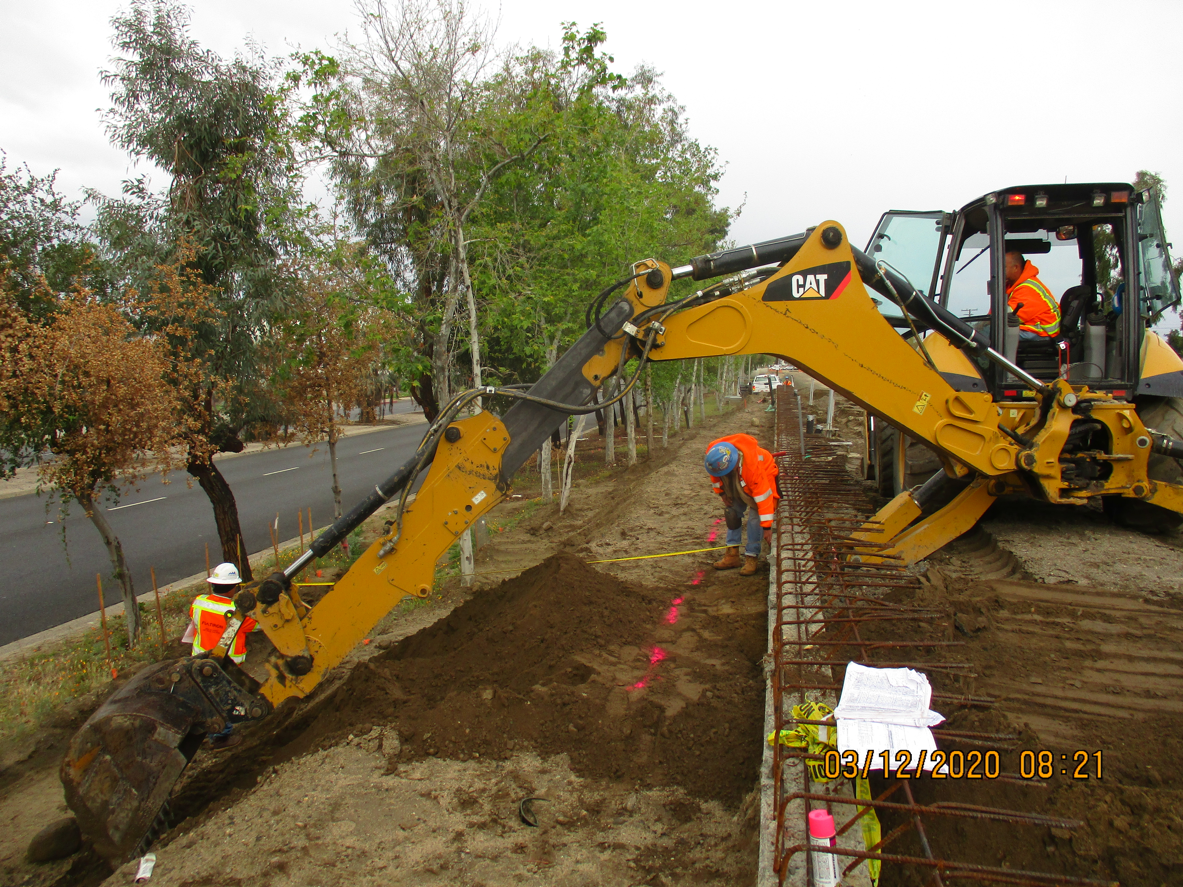 A construction crew uses a backhoe to dig alongside a road, with workers in orange safety vests and hard hats marking the ground near rebar and pink markers.