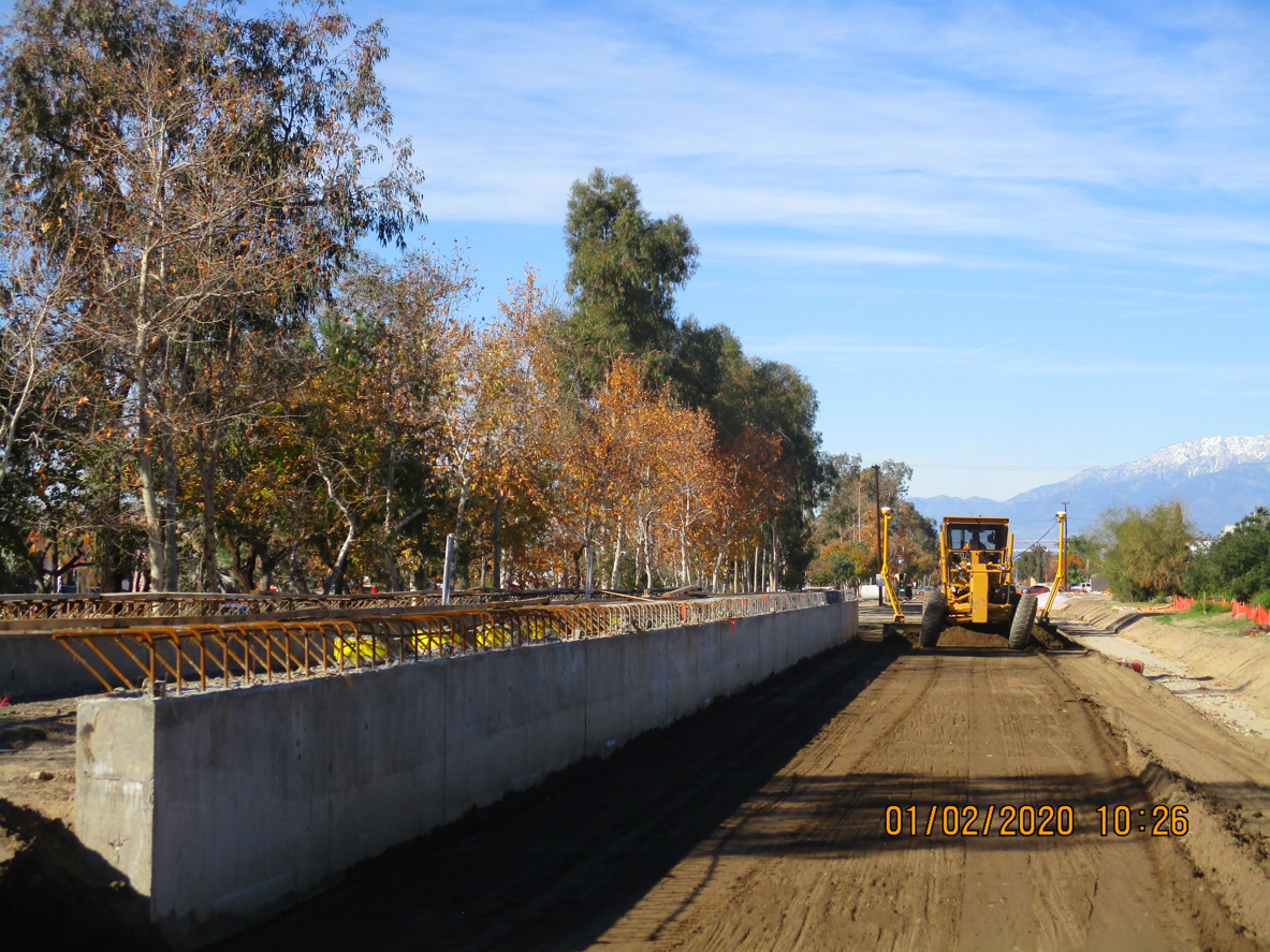 A bulldozer works on a dirt road next to a concrete barrier under clear skies, with trees and distant mountains in the background.