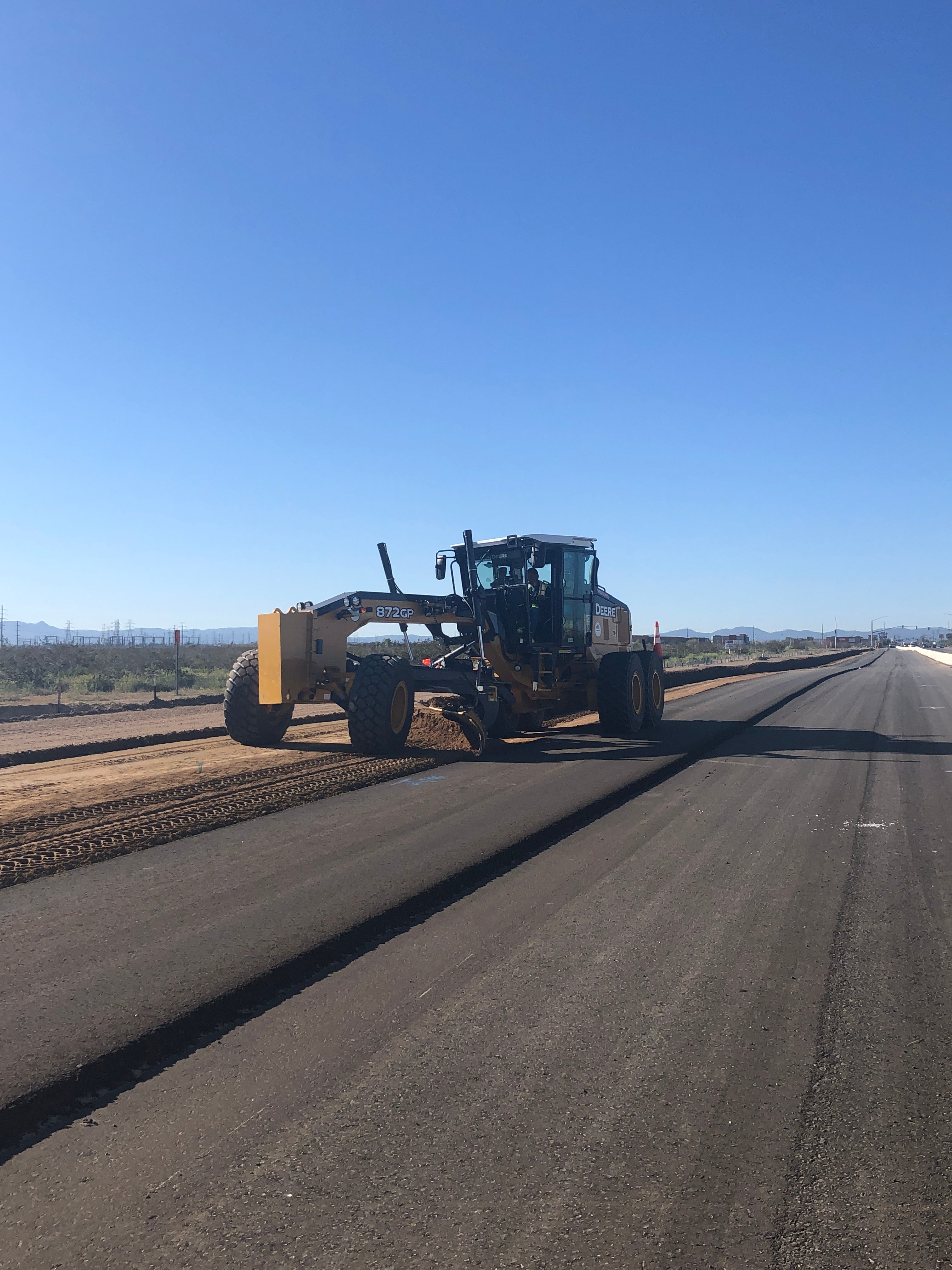 A road grader levels dirt beside a newly paved road under a clear blue sky in a rural area.