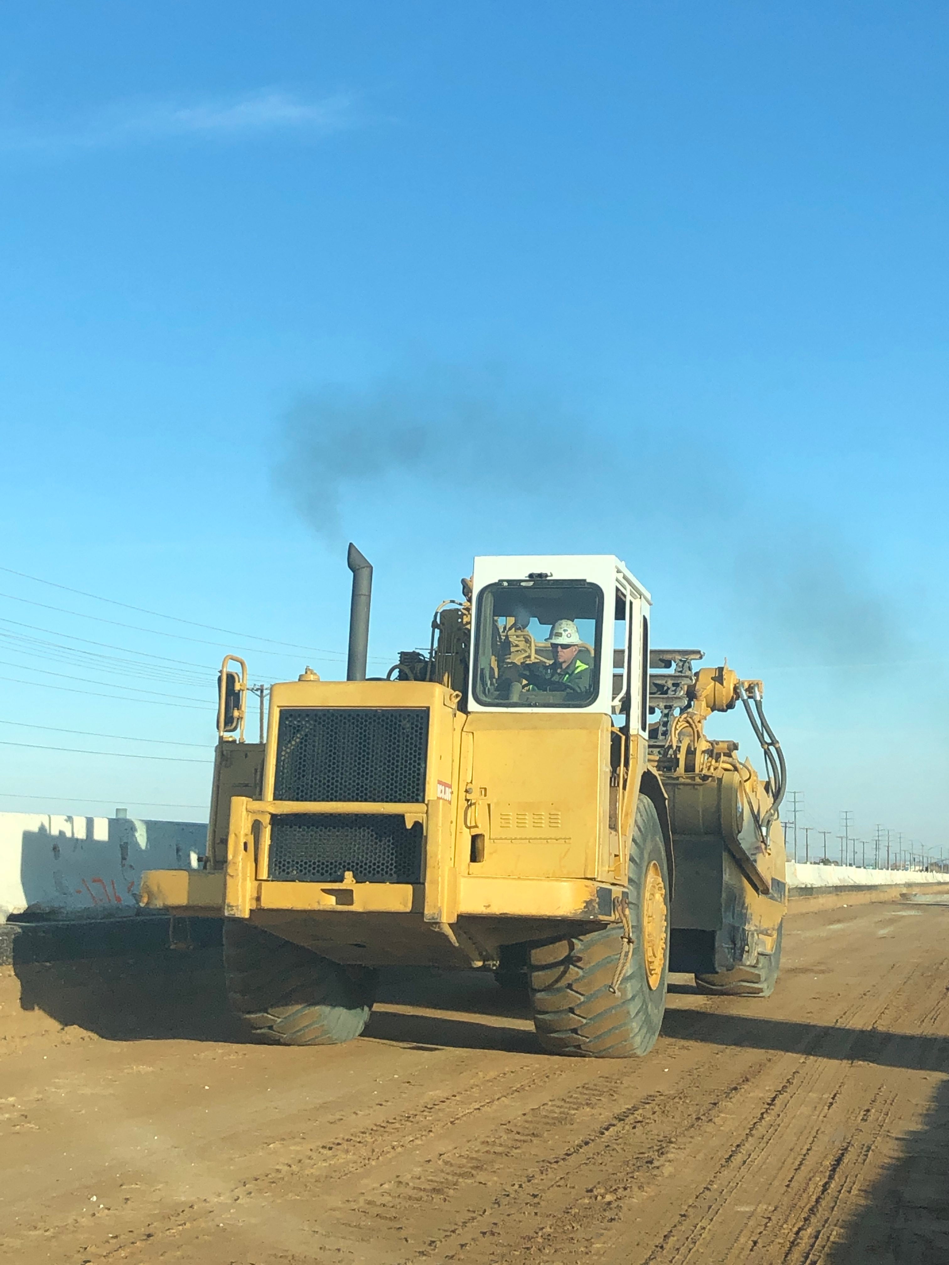 A large yellow construction vehicle drives on a dirt road, emitting black exhaust, with a person visible in the cab under a clear blue sky.