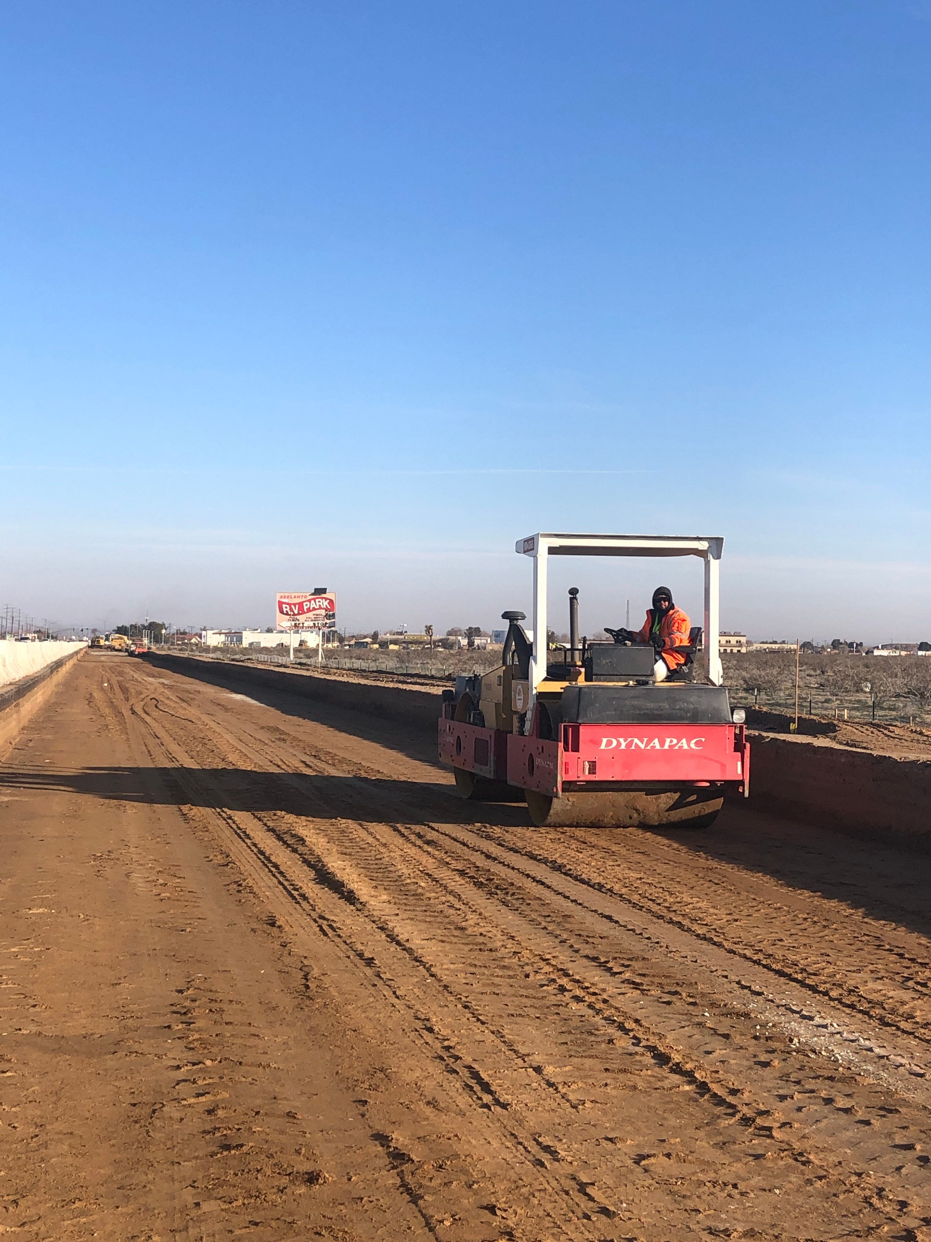 A construction worker operates a Dynapac roller on a dirt road under a clear blue sky, with a Toy Drive sign visible in the background.