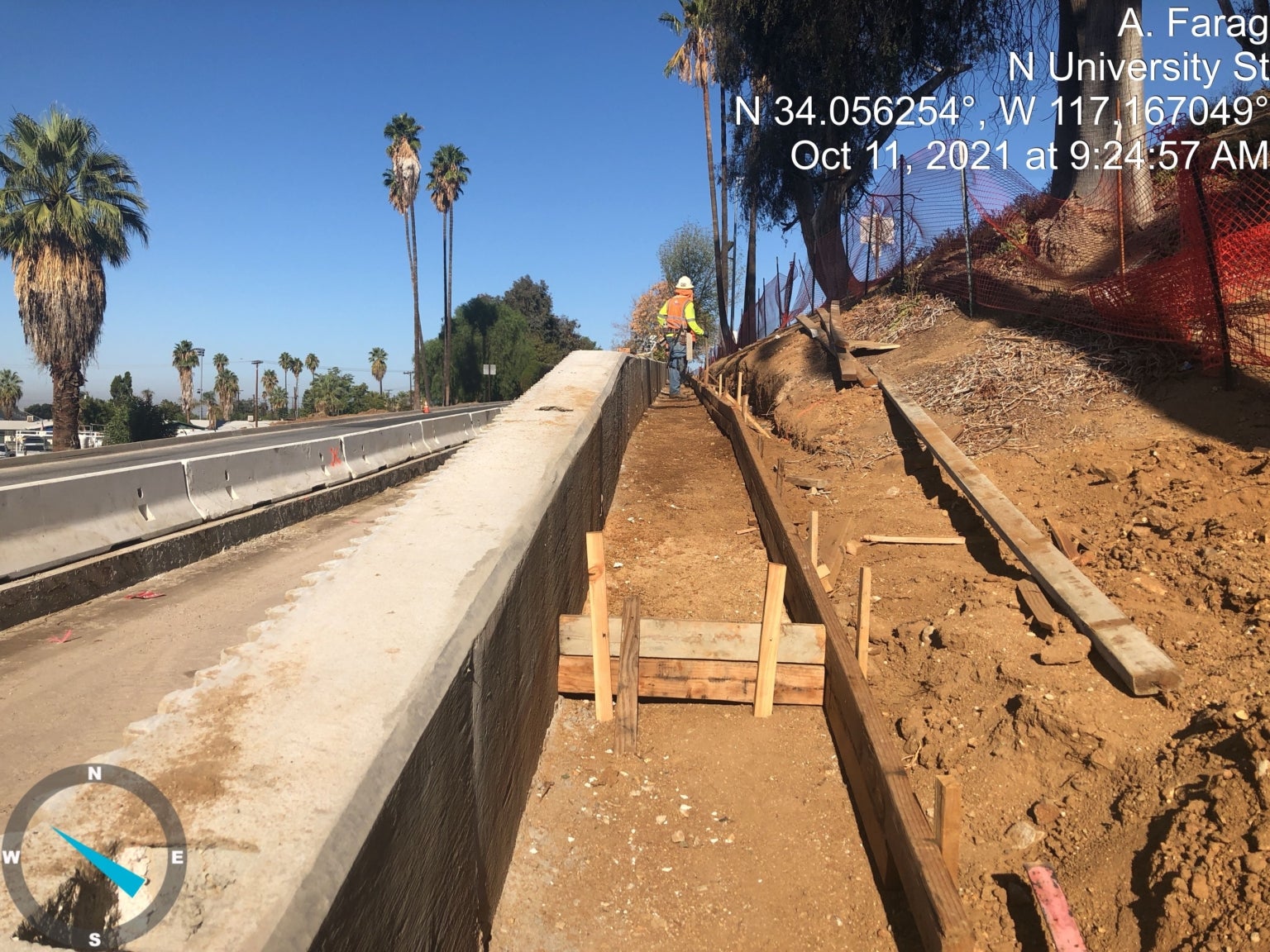 A construction worker stands on a dirt slope next to forms for a concrete curb beside a roadway. Palm trees and construction fencing are visible. Timestamp and coordinates overlay the image.