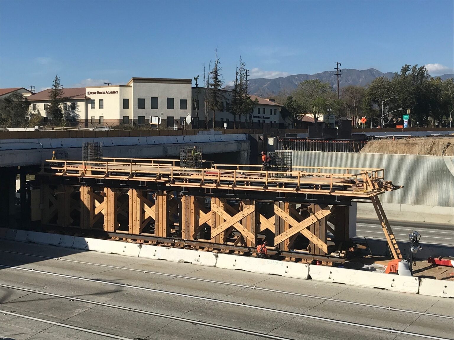 Construction workers stand on scaffolding supporting a bridge over a multi-lane highway, with buildings and mountains visible in the background.