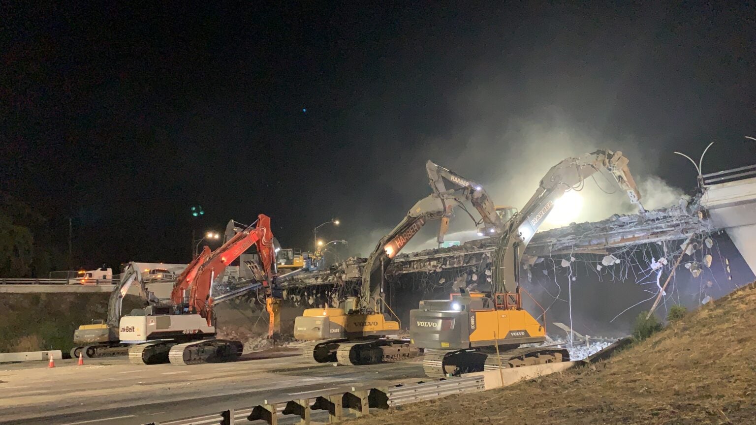 Several excavators demolish a bridge at night, with debris falling and lights illuminating the construction site.