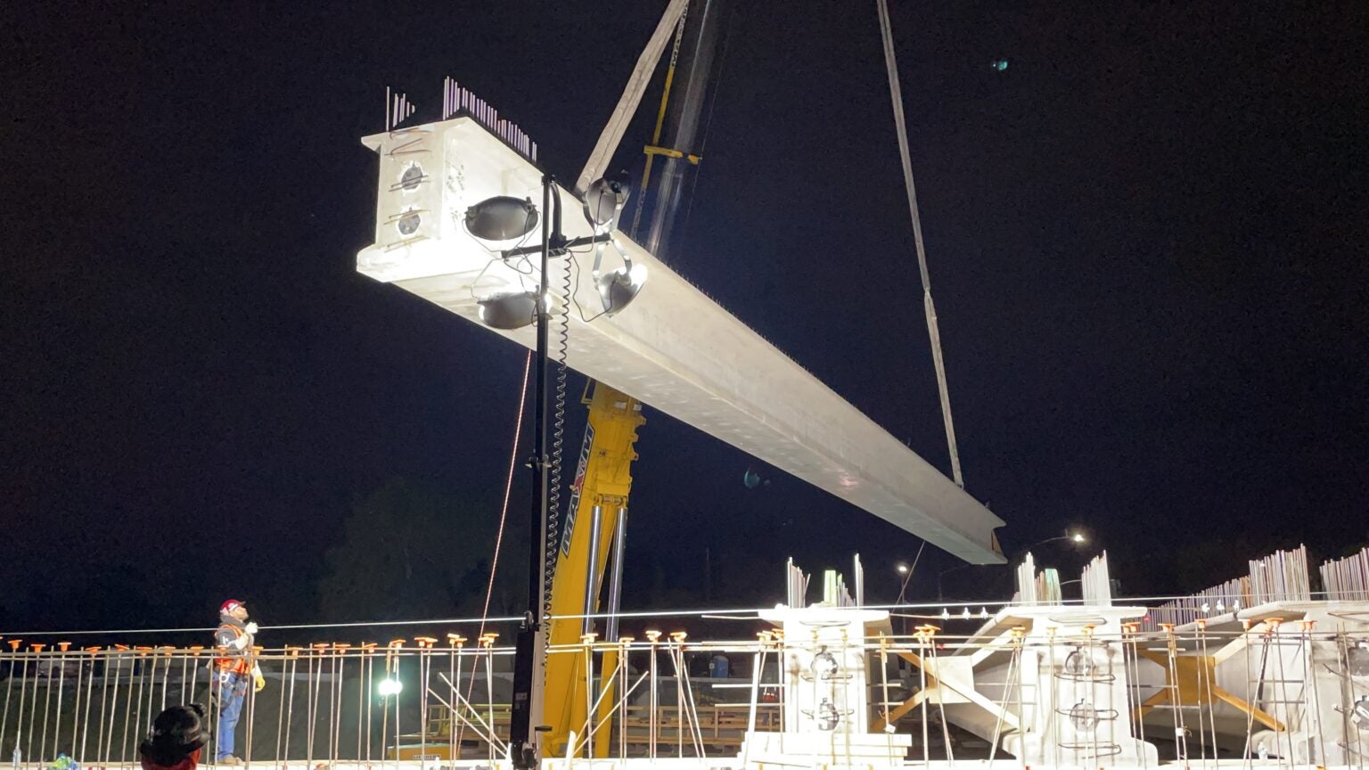 A large concrete beam is being lifted by a crane at a construction site at night, with workers nearby and temporary lighting illuminating the area.