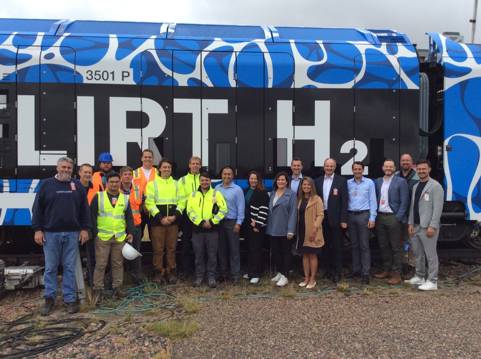 A group of people in business and safety attire stand in front of a blue and black train labeled "FLIRT H₂" outdoors on a cloudy day.