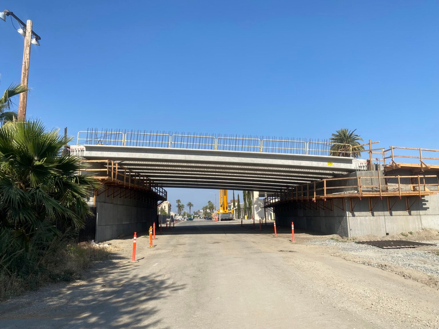 A concrete bridge under construction spans a dirt road, with safety railings and orange traffic cones visible. Palm trees and construction equipment are in the background under a clear sky.