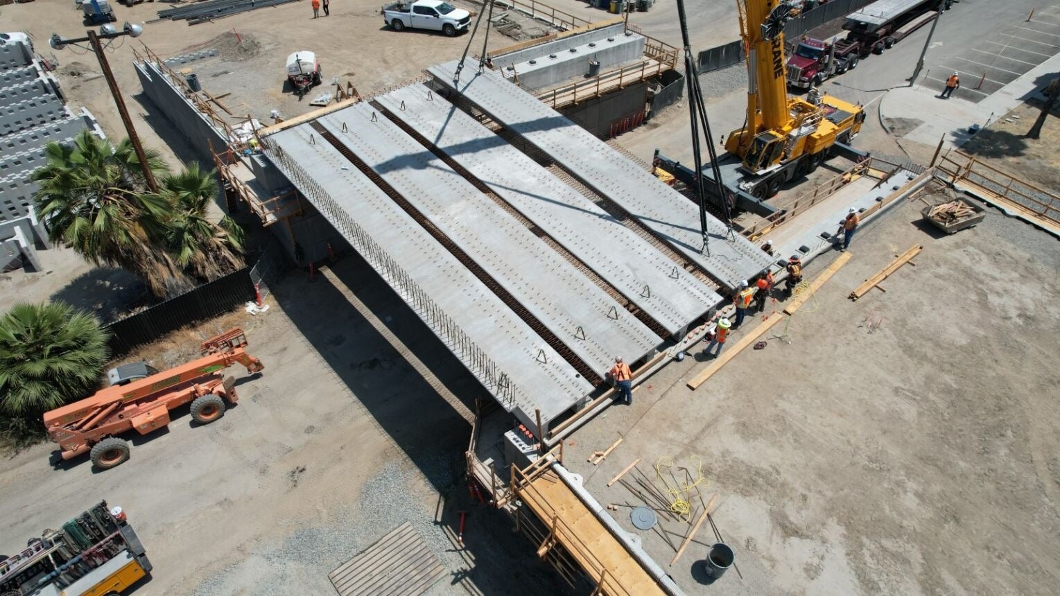 A crane lifts and positions large concrete bridge segments while construction workers guide them into place at a construction site.