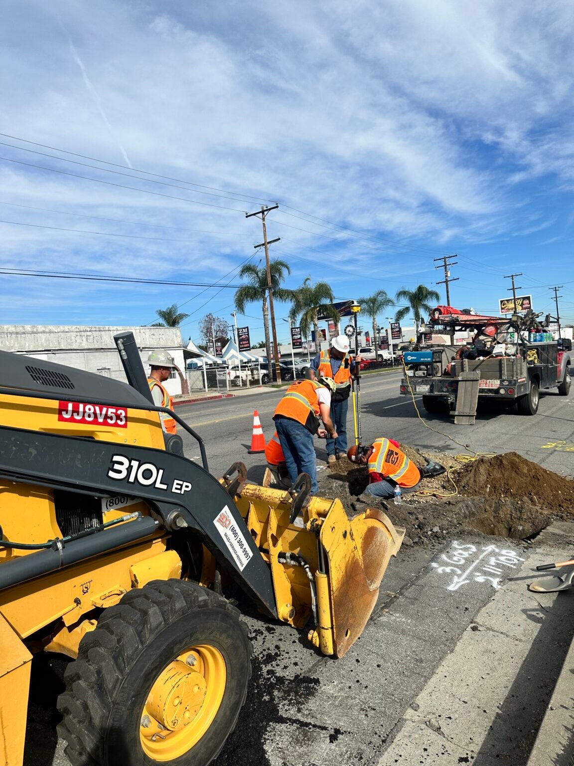Construction workers repair a section of road using a backhoe loader and tools; traffic cones and utility markings are visible on the street.