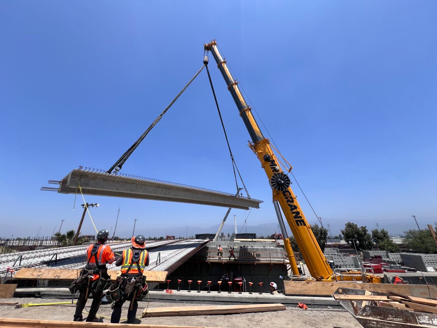 Two construction workers observe as cranes lift a large concrete beam into place over a bridge construction site under a clear blue sky.