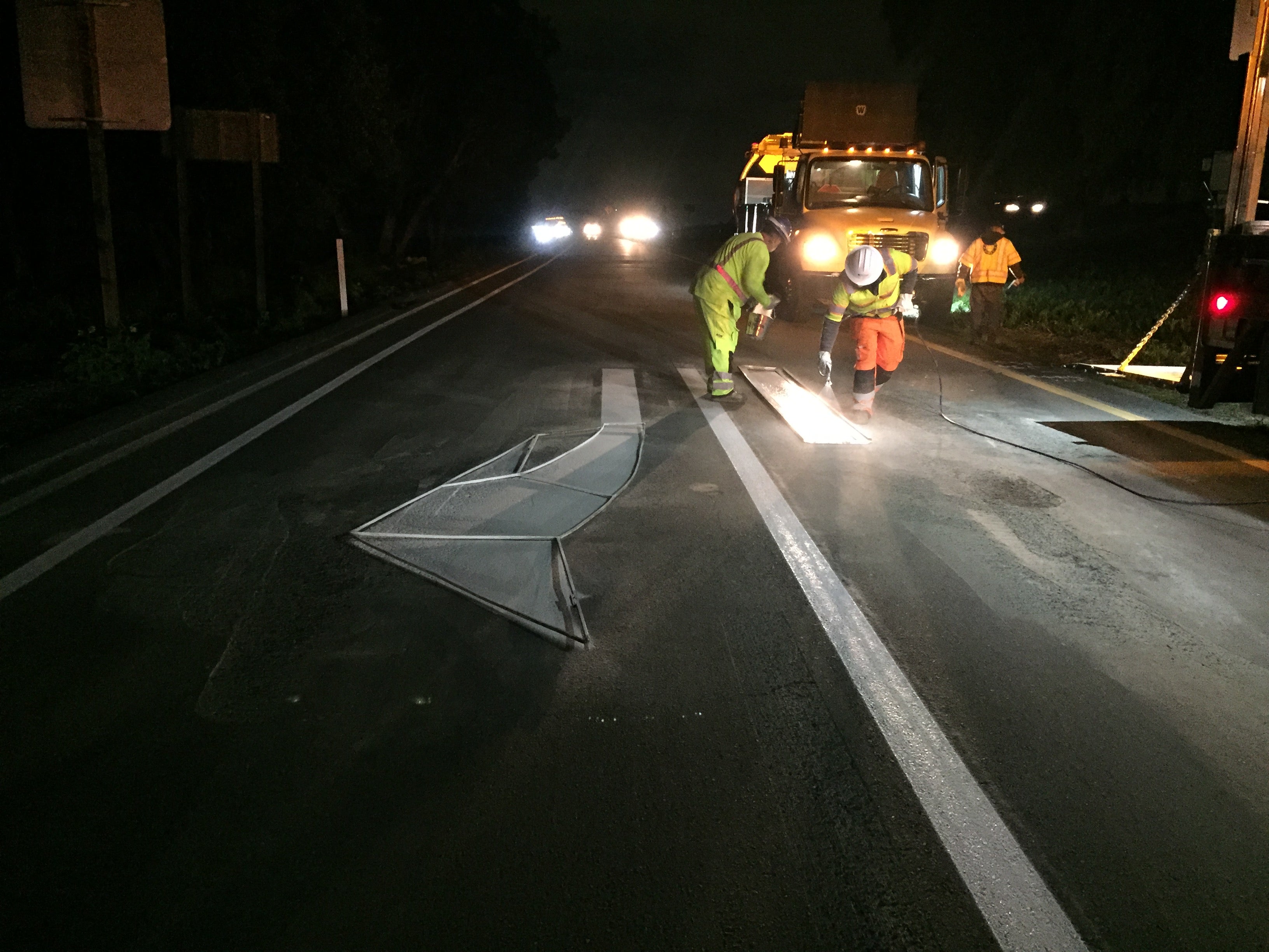 Workers in reflective gear repaint lane markings on a road at night, illuminated by vehicle headlights and a work truck.