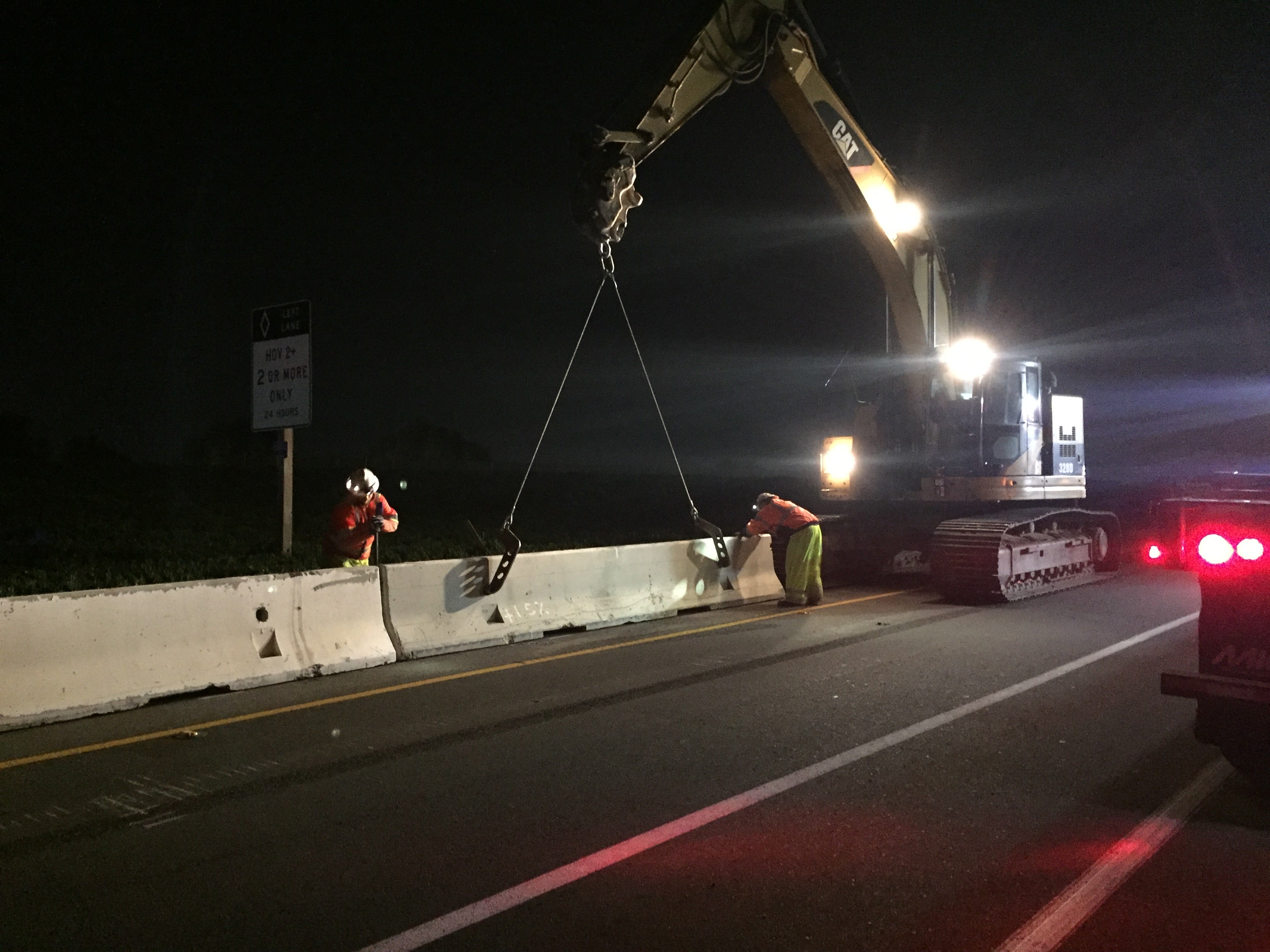 Two construction workers guide a concrete barrier being lifted by a large excavator at night on a road under construction.