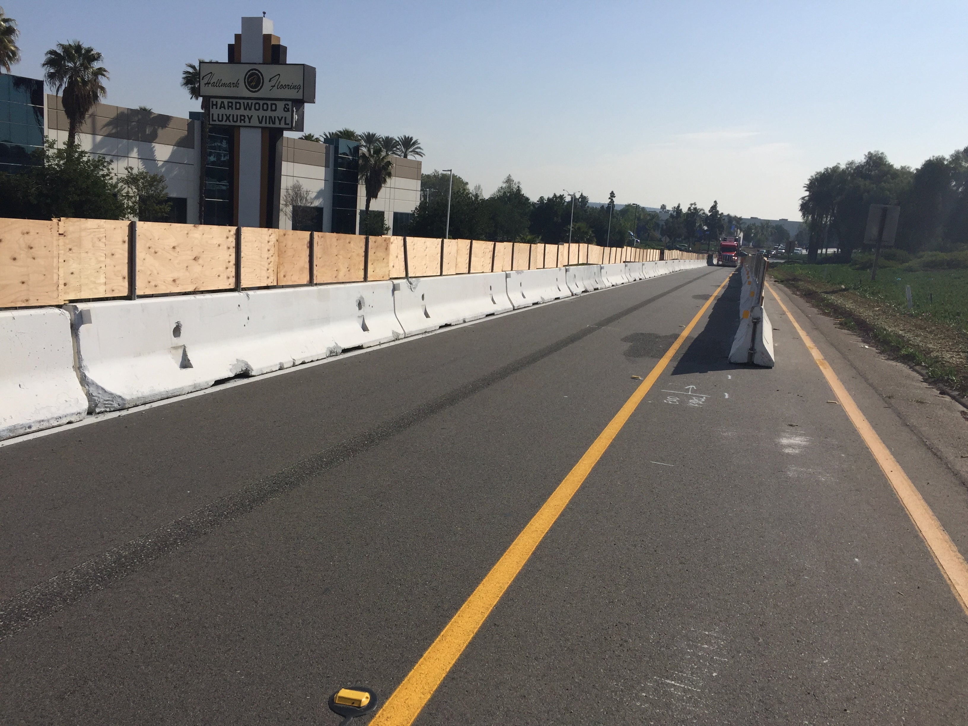 A road is divided by concrete barriers, with wooden fencing on the left. The street is empty, and a building with signage is visible in the background.