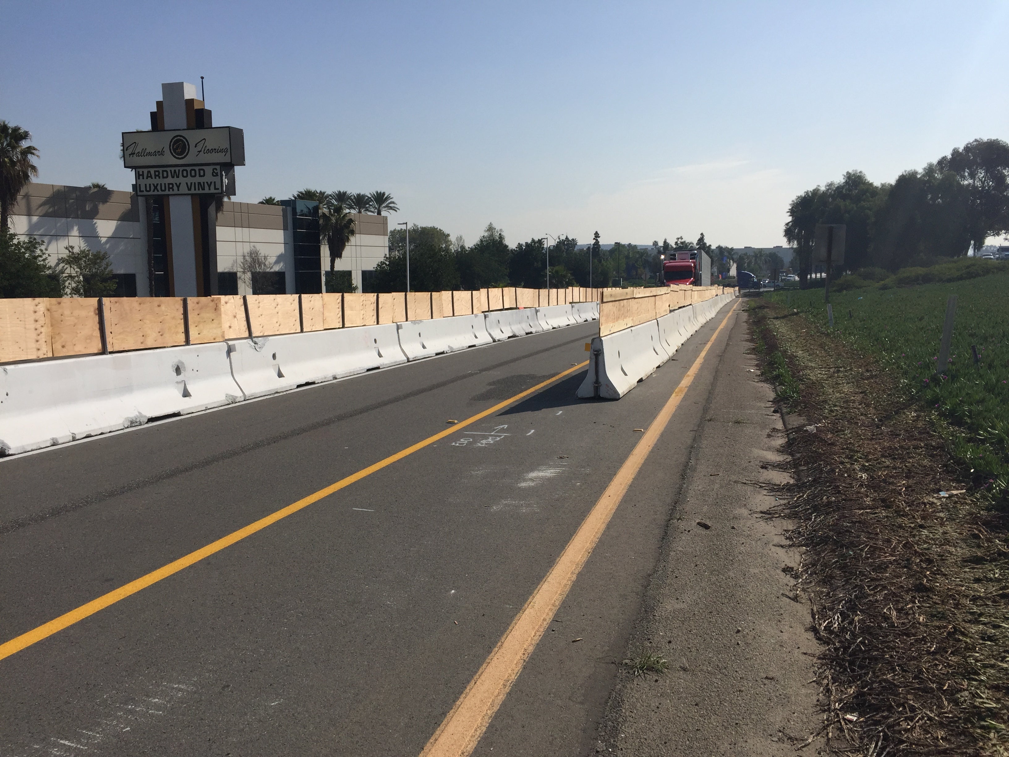 A road is partially closed with concrete and wooden barriers, blocking one lane; a truck is visible in the distance, and a sign for a flooring store is at left.