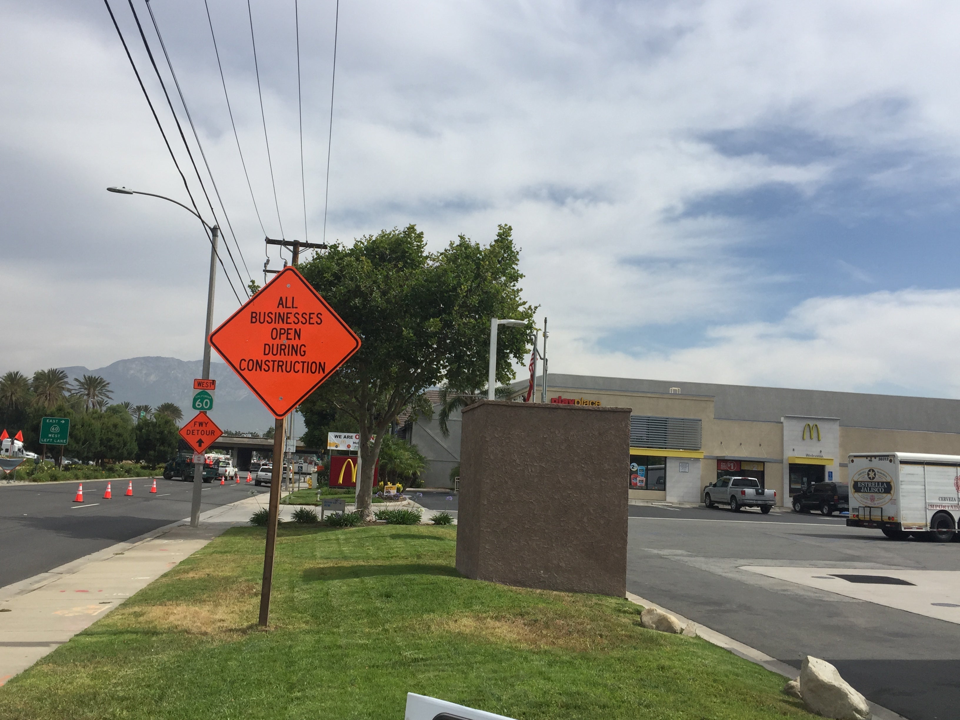Orange sign on a grassy median reads "All businesses open during construction." Fast food restaurants and cars are visible in the background.
