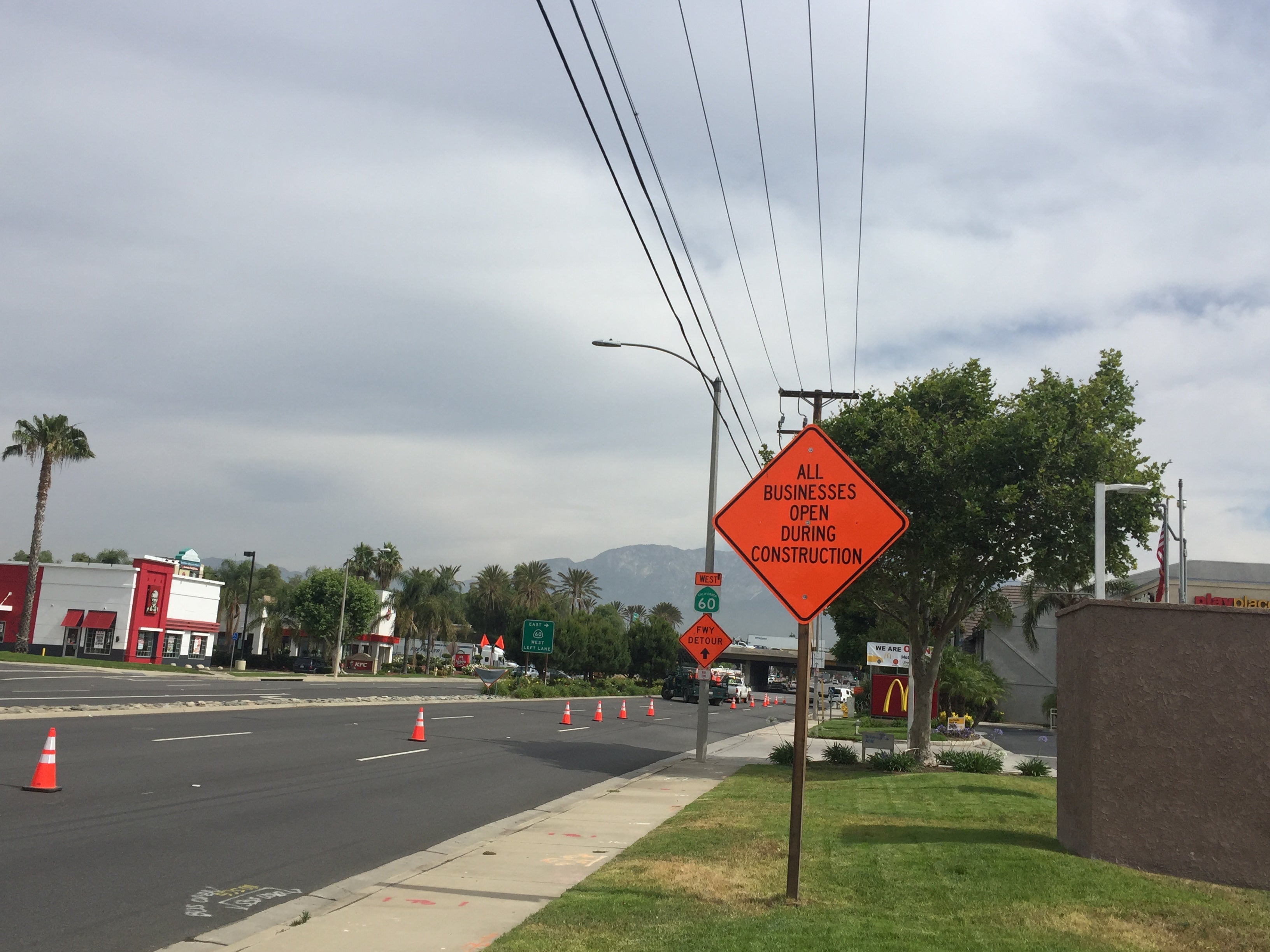A street with traffic cones along the sidewalk and a sign reading "All businesses open during construction"; businesses and a McDonald's are visible in the background.