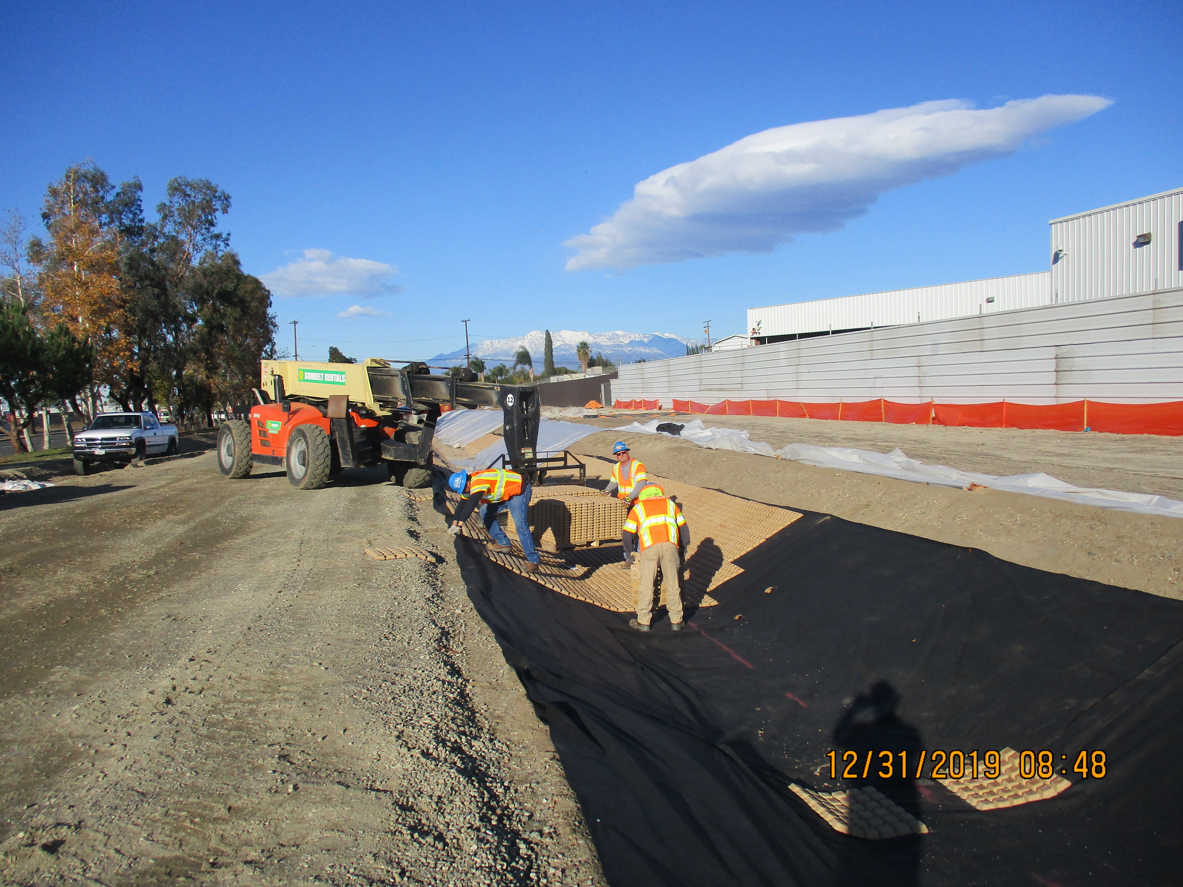 Construction workers in safety gear install a large black liner on a dirt site next to a building, with machinery and vehicles present under a clear sky.