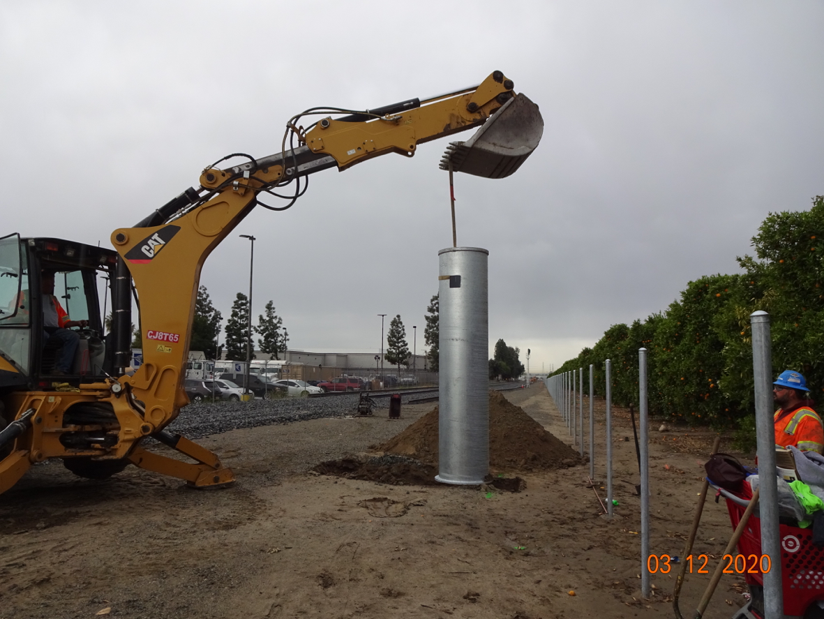 A backhoe lifts a large cylindrical metal post near a fenced area; workers in safety gear observe the installation on a cloudy day.