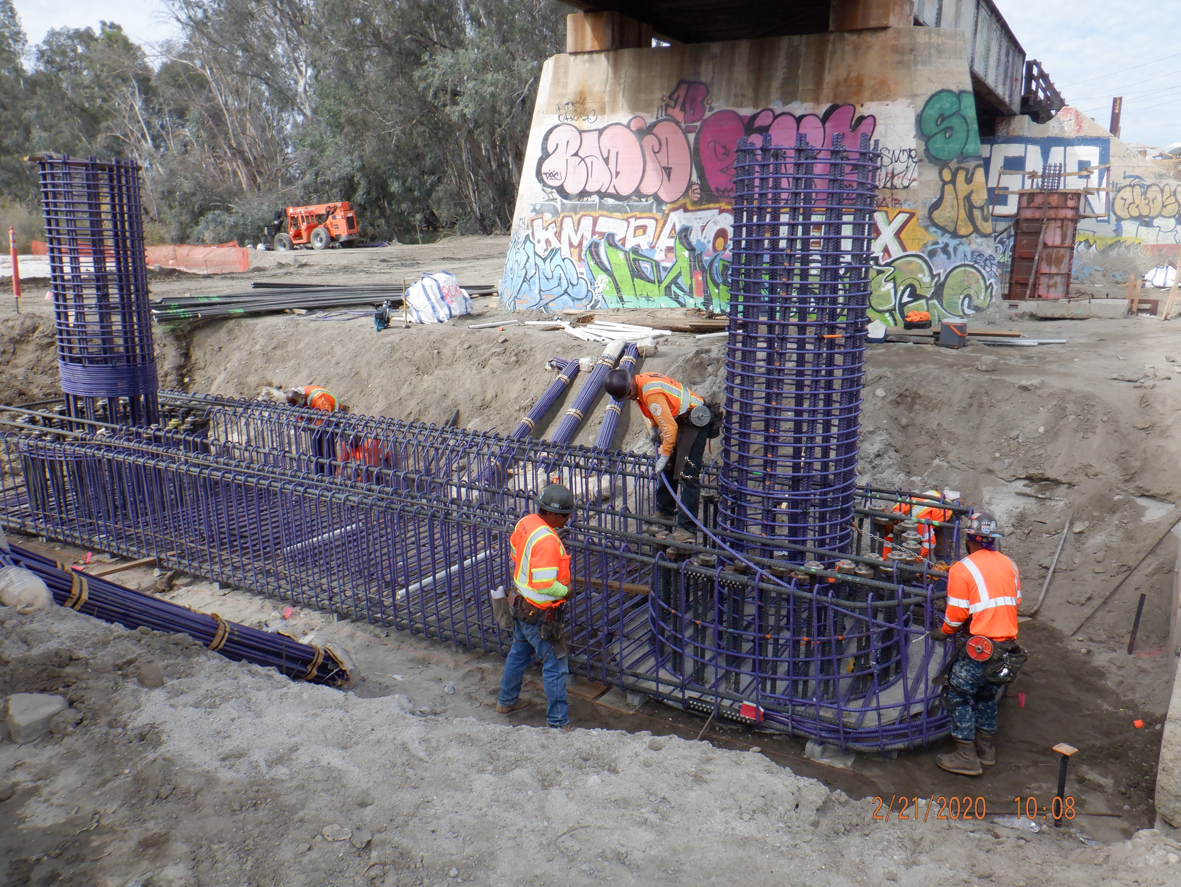 Construction workers assemble steel rebar cages for a bridge foundation near a graffiti-covered overpass, surrounded by construction equipment and materials.