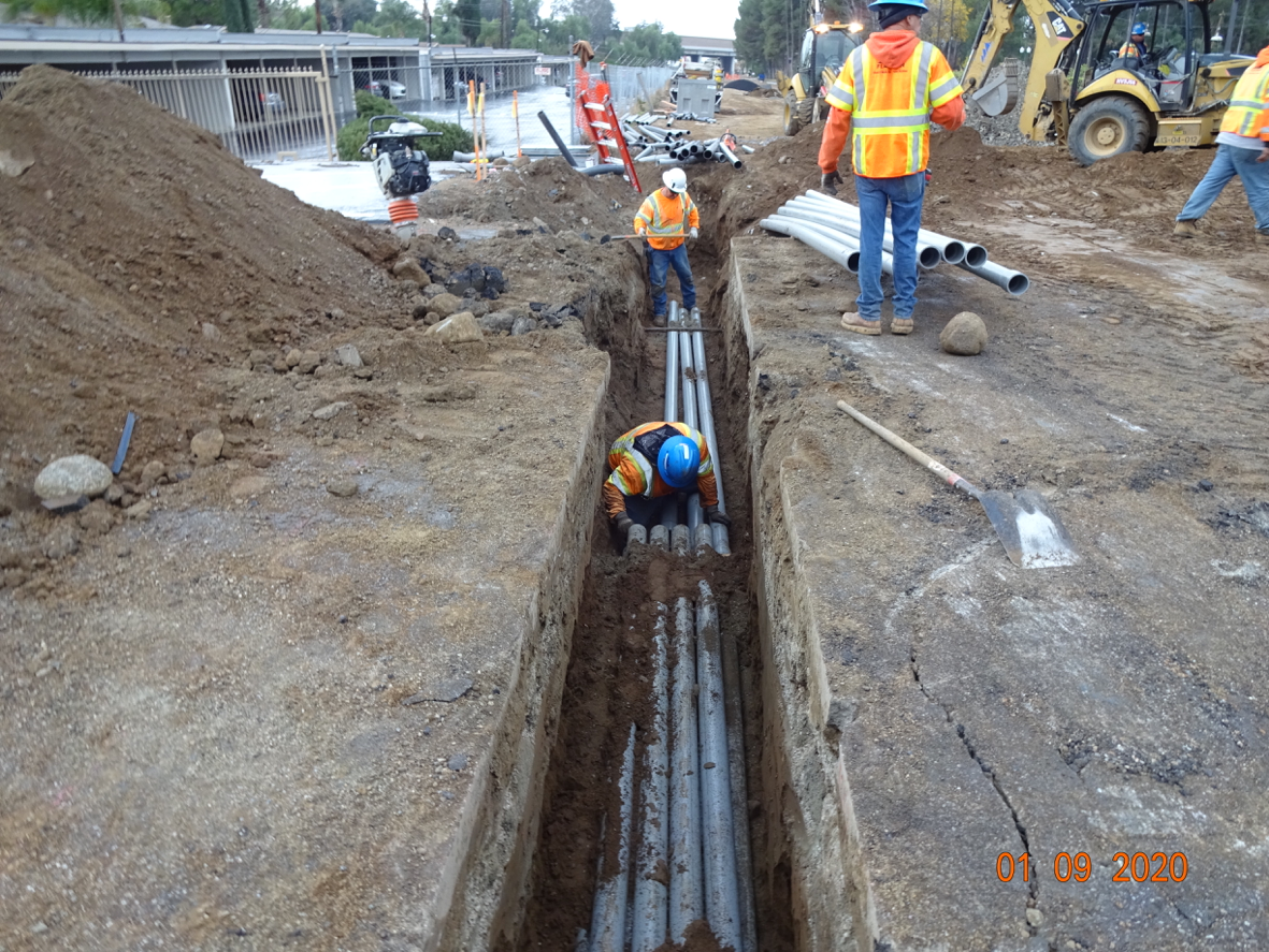 Construction workers in safety gear install and arrange large pipes in a trench at a roadside work site, with equipment and barriers visible in the background.