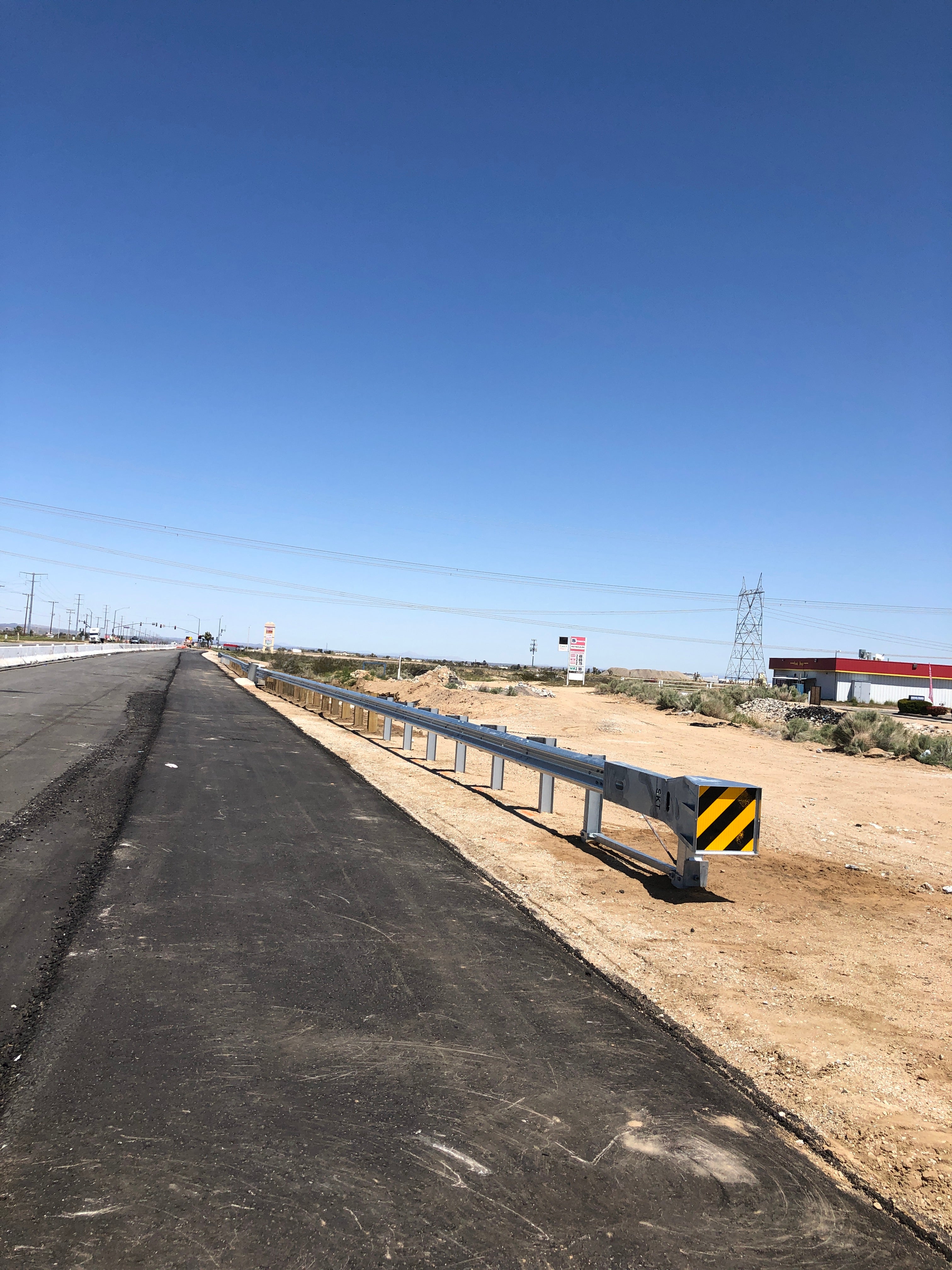 A newly paved road with a metal guardrail along the right side, set in a dry, open area with a clear blue sky overhead.
