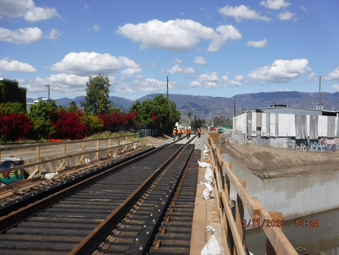 Railroad tracks under construction with several workers in safety gear in the distance; mountains and clouds in the background.