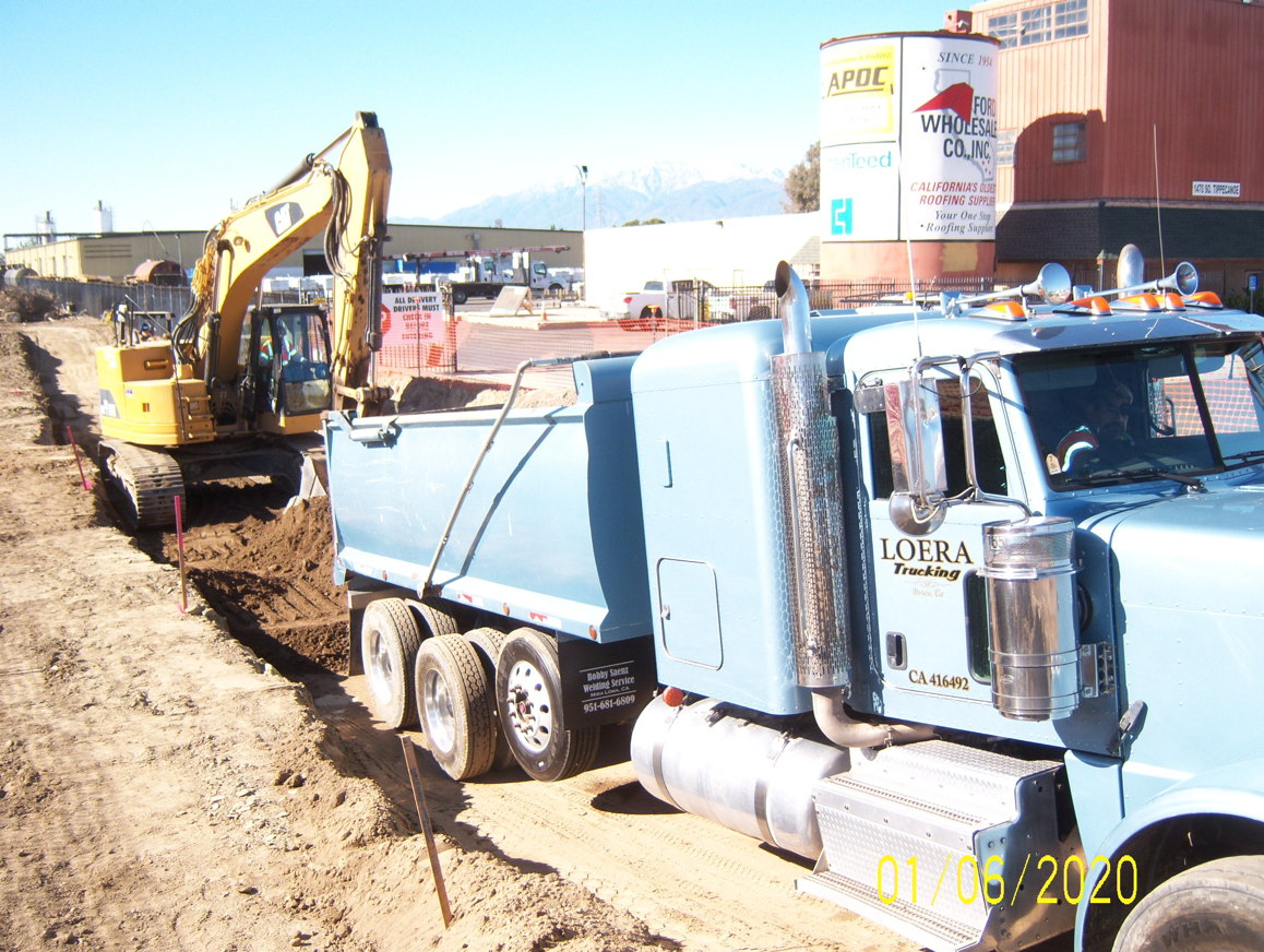 An excavator loads soil into a blue dump truck at a construction site with industrial buildings and equipment in the background.