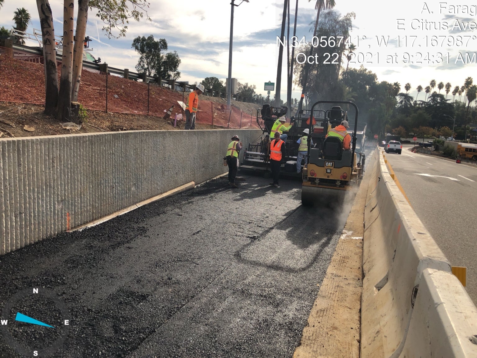 Construction workers pave a new asphalt road beside a concrete barrier; machinery and safety cones are visible. Timestamp, coordinates, and location text overlay the image.