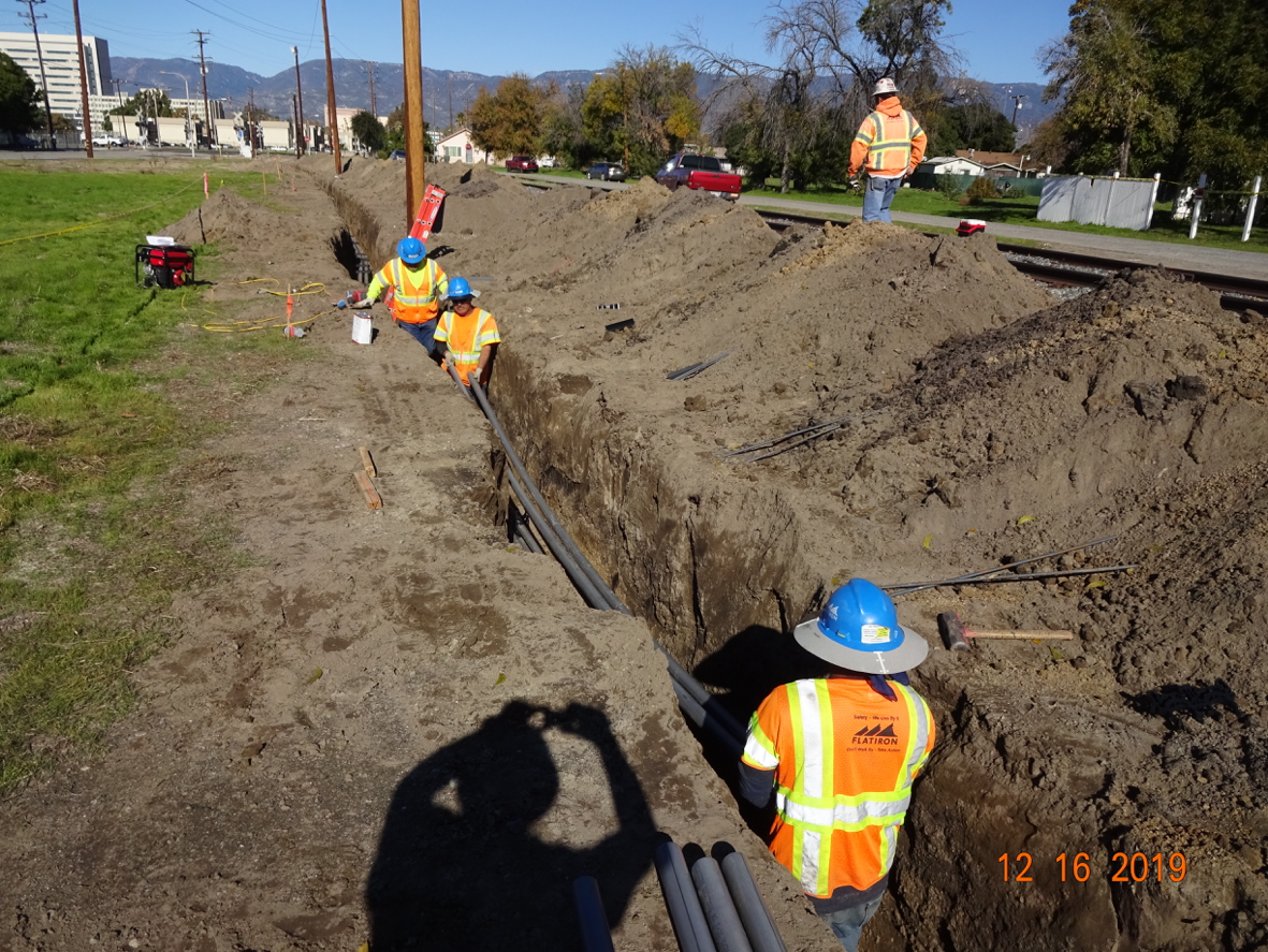 Four construction workers install conduit pipes in a trench alongside a road and railroad tracks on a sunny day. Large piles of dirt are visible beside the trench.