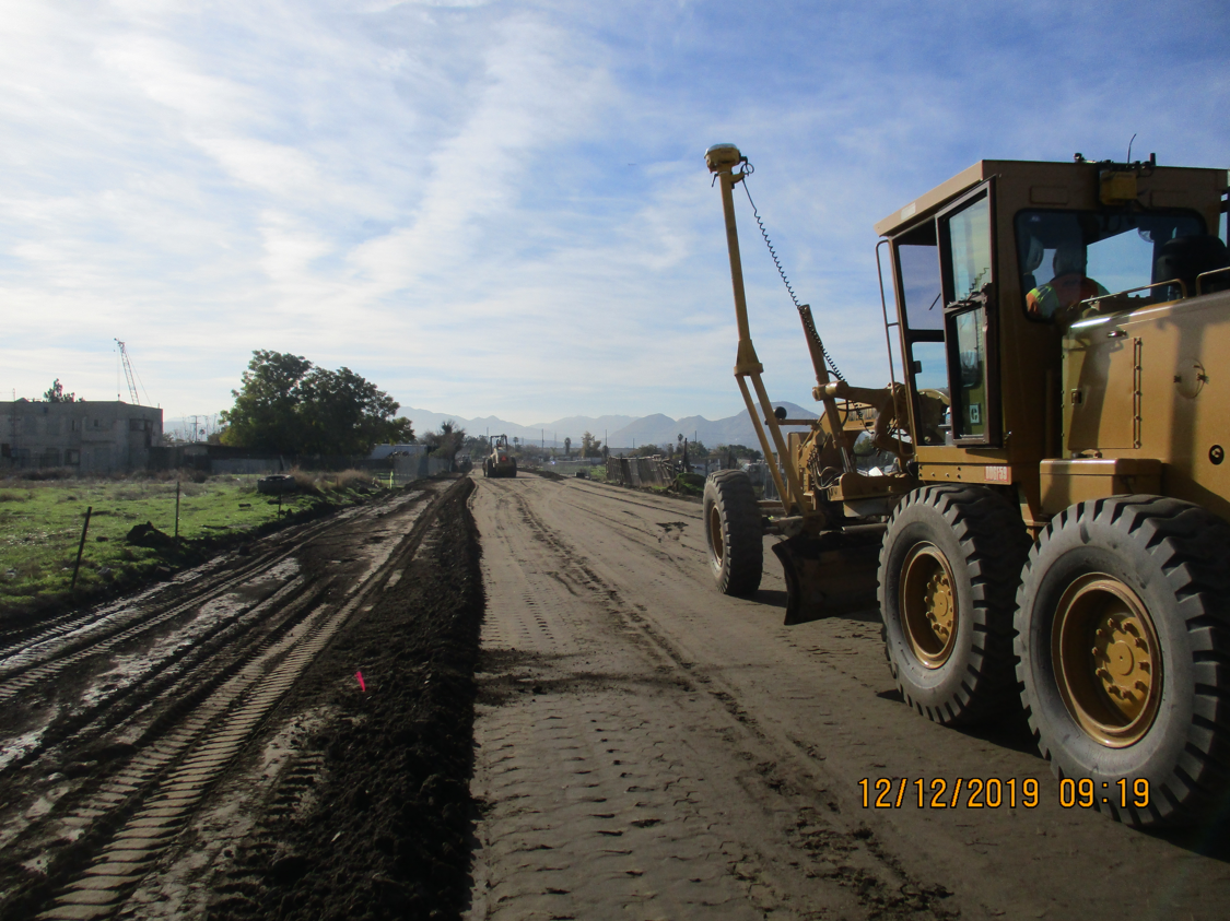 A construction grader levels a dirt road under a partly cloudy sky, with another vehicle visible in the distance; photo dated 12/12/2019.