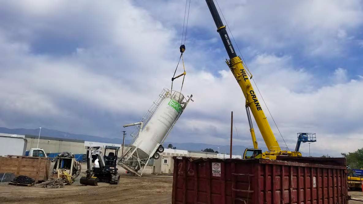 A crane lifts a large white silo, tilting it upright at a construction site with scattered equipment and containers under a cloudy sky.