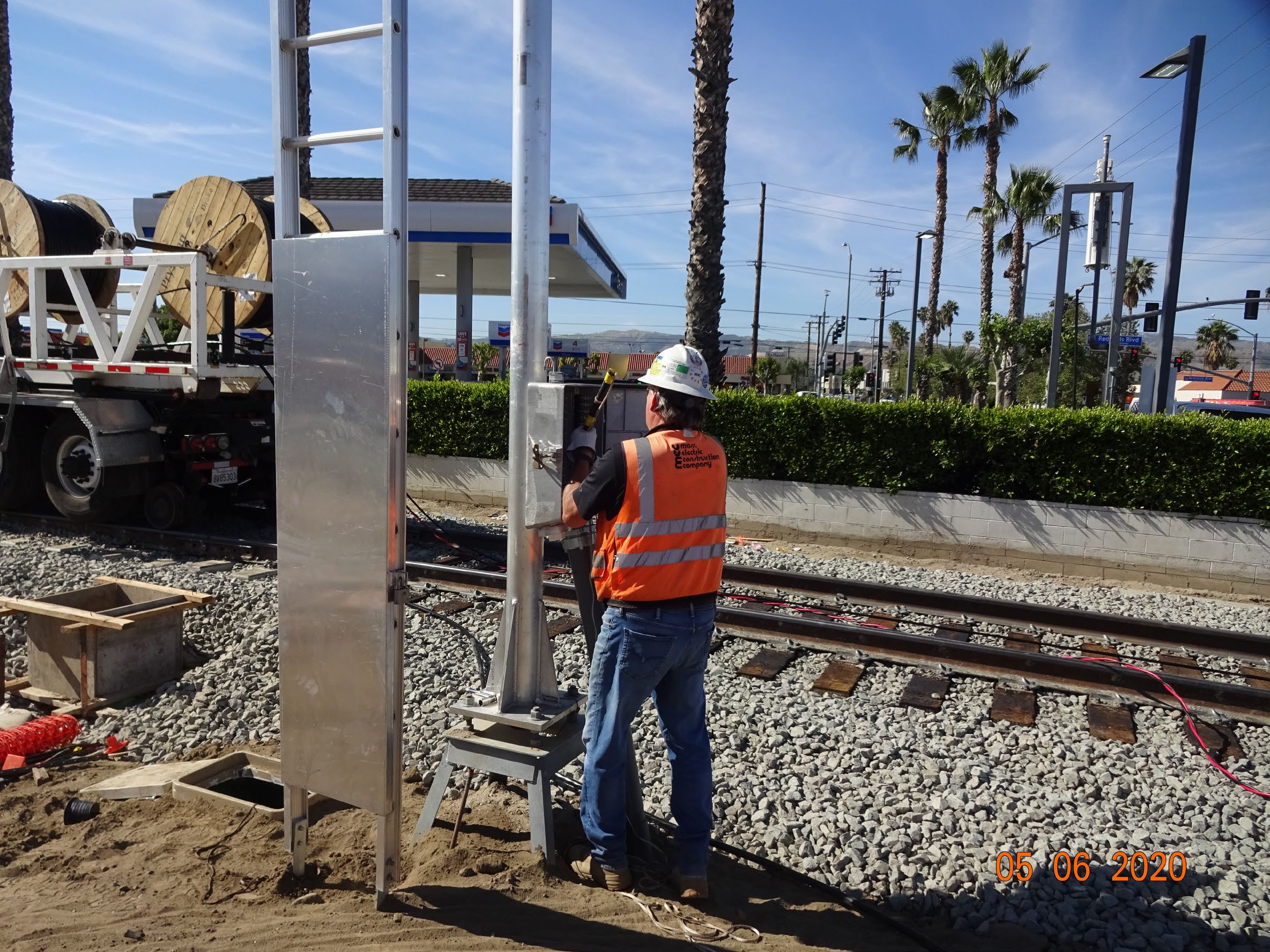 A construction worker in a safety vest and helmet installs a metal structure beside railroad tracks on a sunny day.