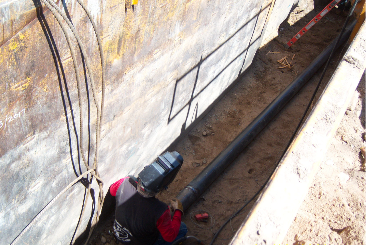 A worker wearing a welding helmet is welding a black pipeline in a trench with metal walls and exposed soil.