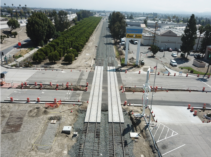 A railroad track crosses a street under construction, with traffic cones and barriers. Commercial buildings and parking lots are visible on both sides of the road.